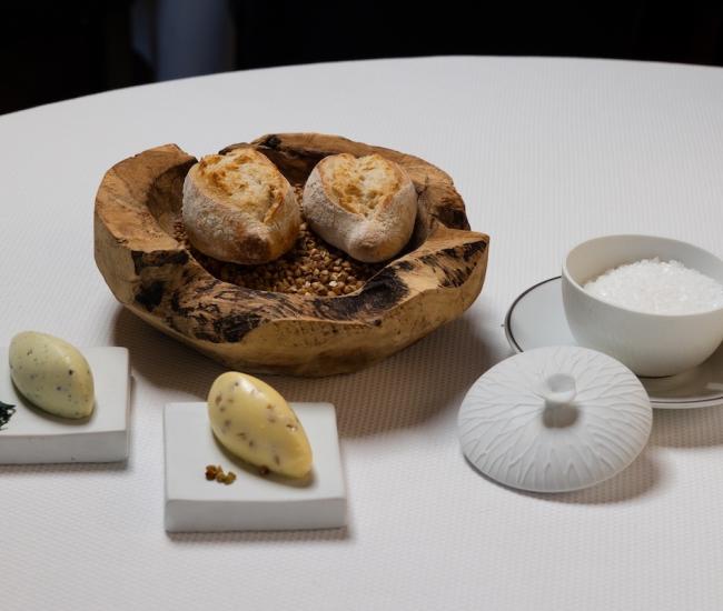 Table de restaurant élégante avec pain maison dans un bol en bois, beurre aux algues et au sarrasin, verres et assiette dressée dans une ambiance chaleureuse.